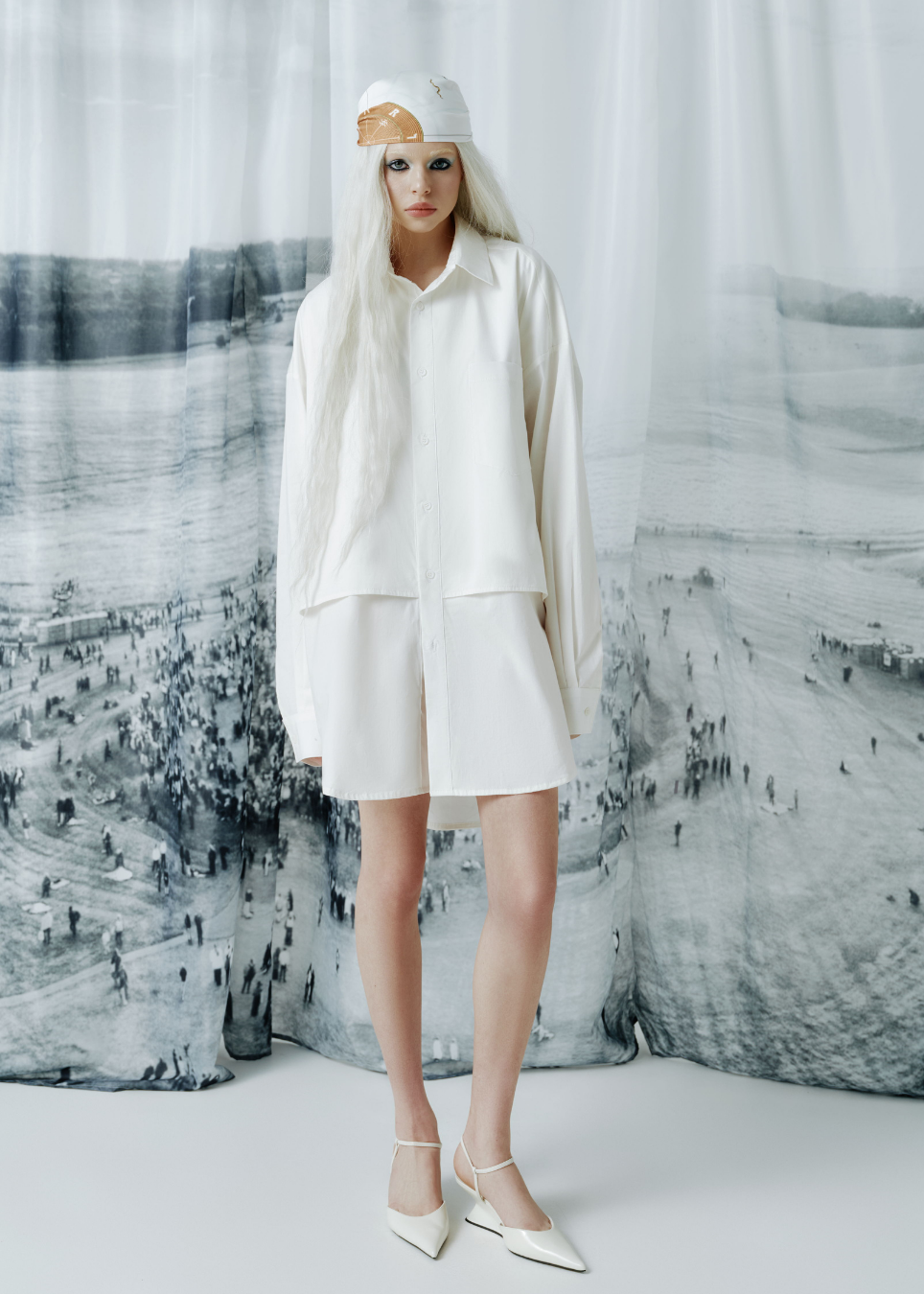 A model wearing a white oversized long-sleeve shirt, matching shorts, and white pointed-toe heels, standing in front of a large black and white landscape photograph backdrop.