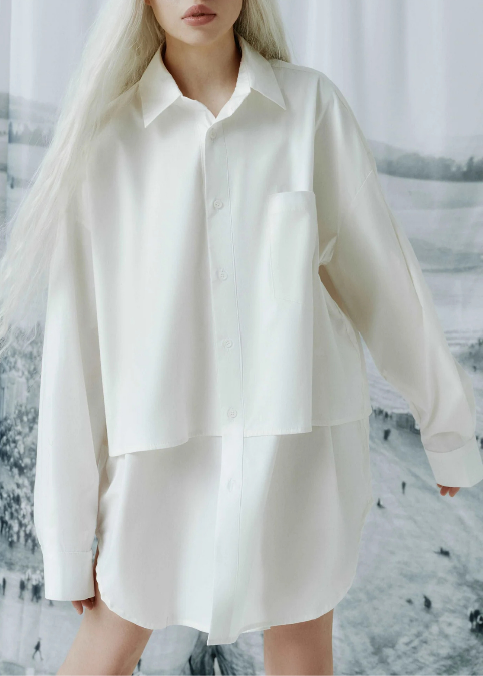 A woman wearing an oversized white long-sleeve button-up shirt dress with a chest pocket, shown against a snowy outdoor backdrop.
