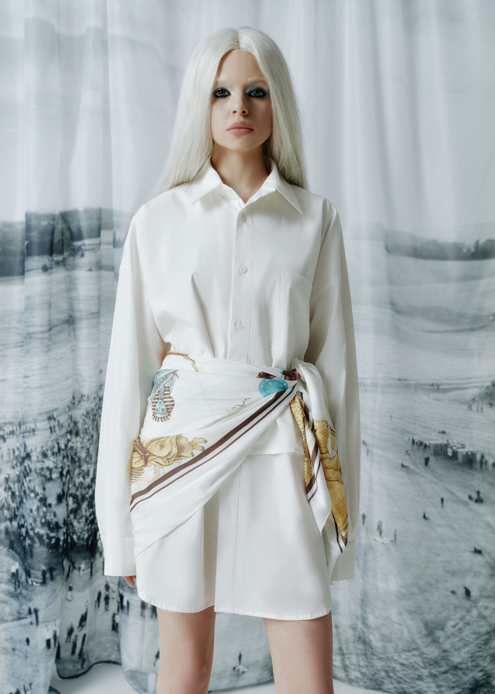 A model with long platinum blonde hair stands in front of a scenic, snowy landscape backdrop, wearing a white long-sleeve button-up shirt and a matching white wrap skirt featuring a gold and blue patterned scarf tied at the waist.