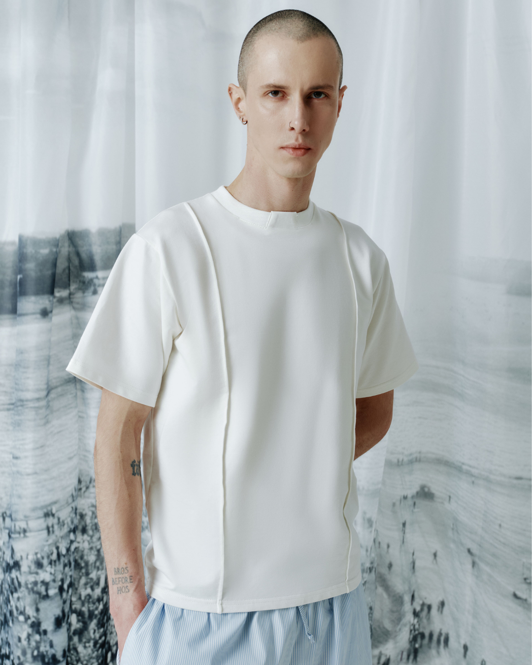 A man wearing a white short-sleeve t-shirt with unique architectural side seams and light blue shorts, standing in front of a white curtain backdrop with a faint landscape print.