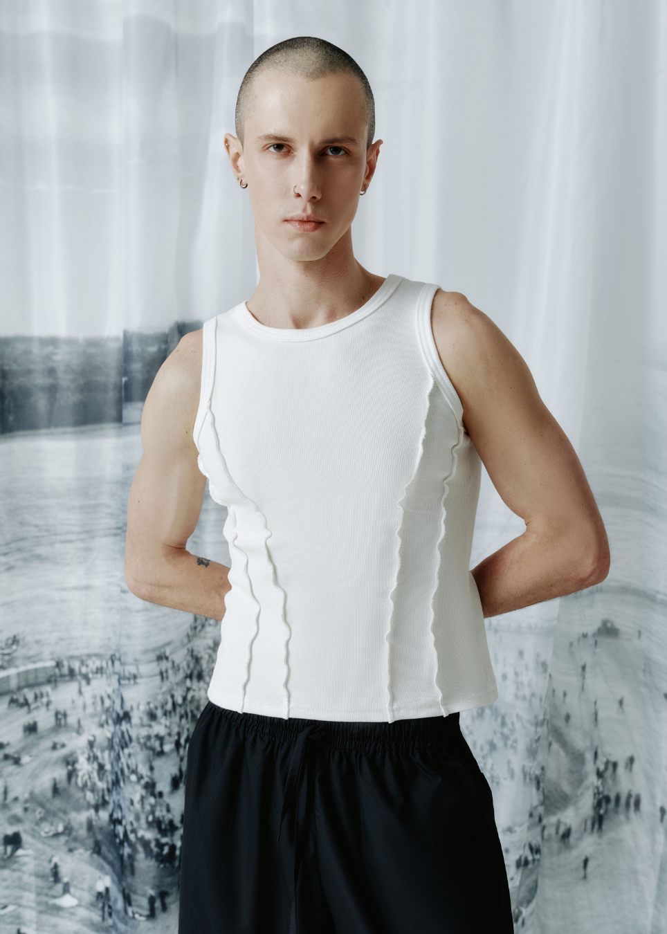 A man wearing a white ribbed sleeveless tank top with vertical pleated details and black shorts, standing against a white curtain backdrop with a black-and-white cityscape print.