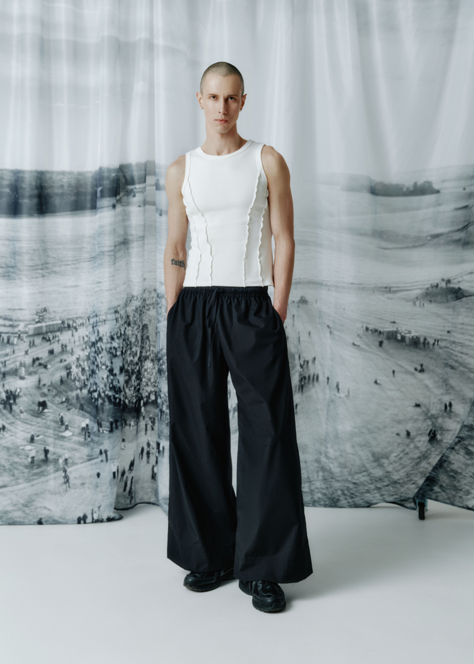 A model stands in a studio wearing a white sleeveless corset-style top with vertical ruffles and black wide-leg trousers, posed in front of a large black-and-white scenic backdrop.