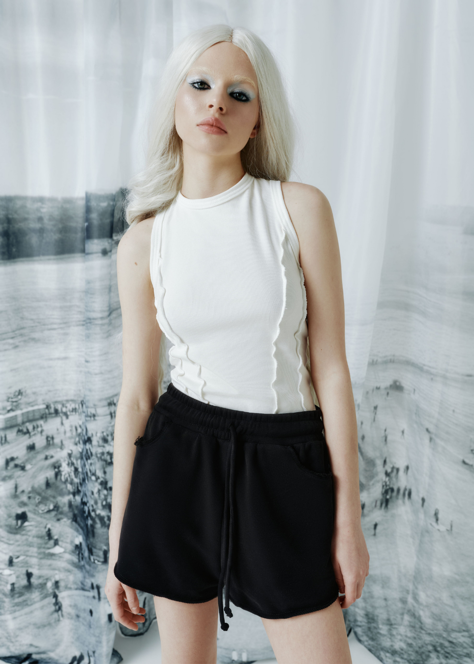 A model with platinum blonde hair wearing a white sleeveless ruffled top and black shorts, standing in front of a white curtain backdrop with a black and white cityscape print.