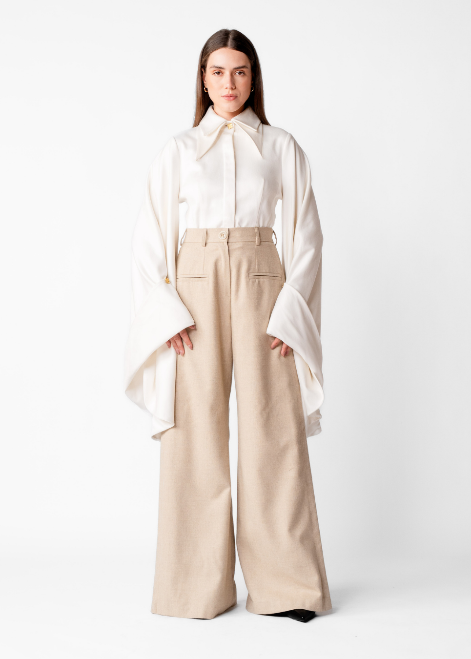 A model stands against a plain white studio background wearing a white long-sleeve blouse with dramatic wide bell sleeves and a high neck, paired with high-waisted beige wide-leg trousers.