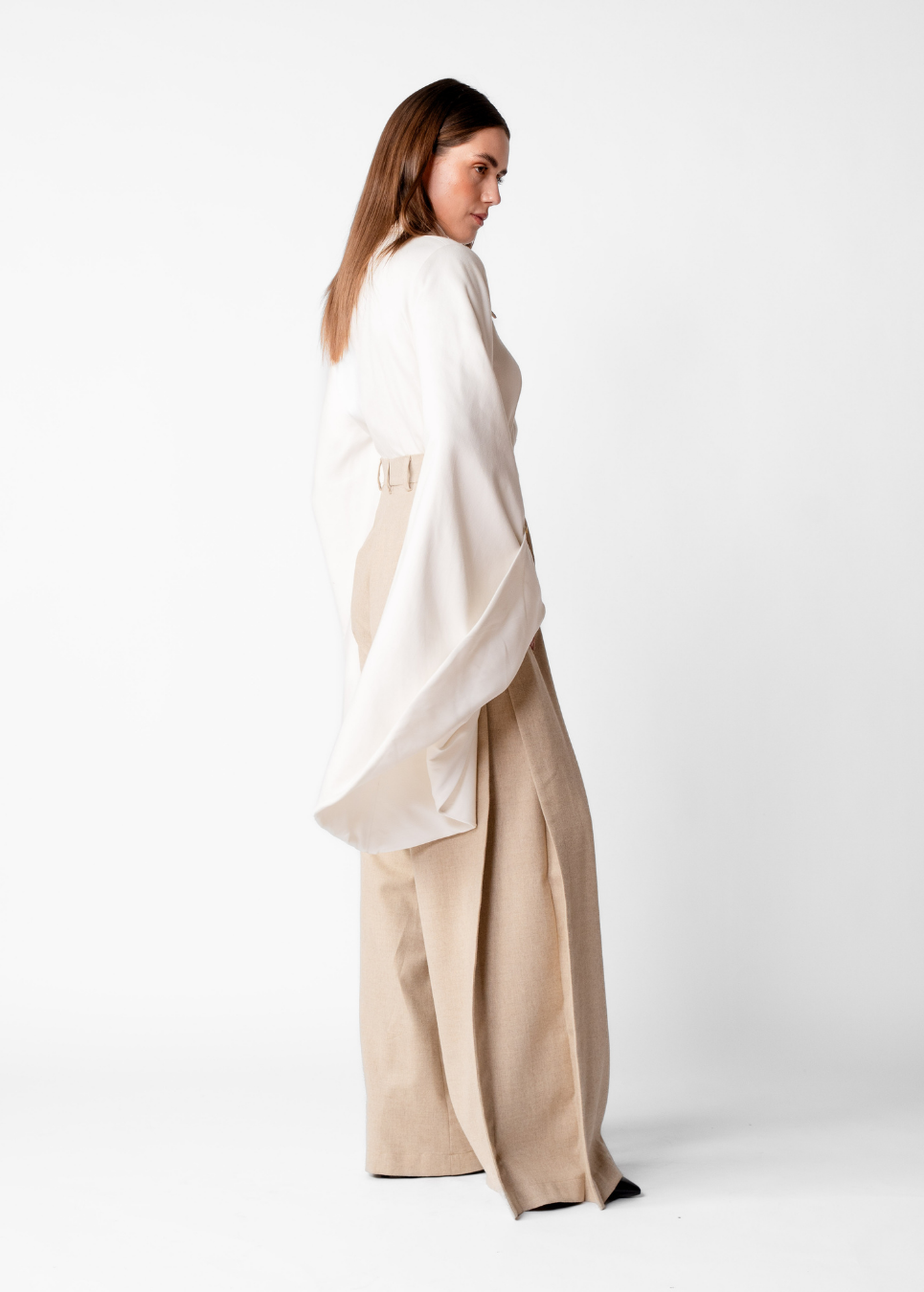 A woman modeling a two-piece outfit consisting of a loose white draped top and wide-leg beige trousers, photographed against a seamless white studio background.