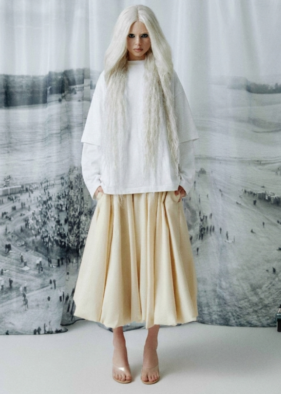A model with long platinum blonde hair stands in front of a large black and white landscape photograph backdrop, wearing a white long-sleeve top and a pleated sand-colored midi skirt.