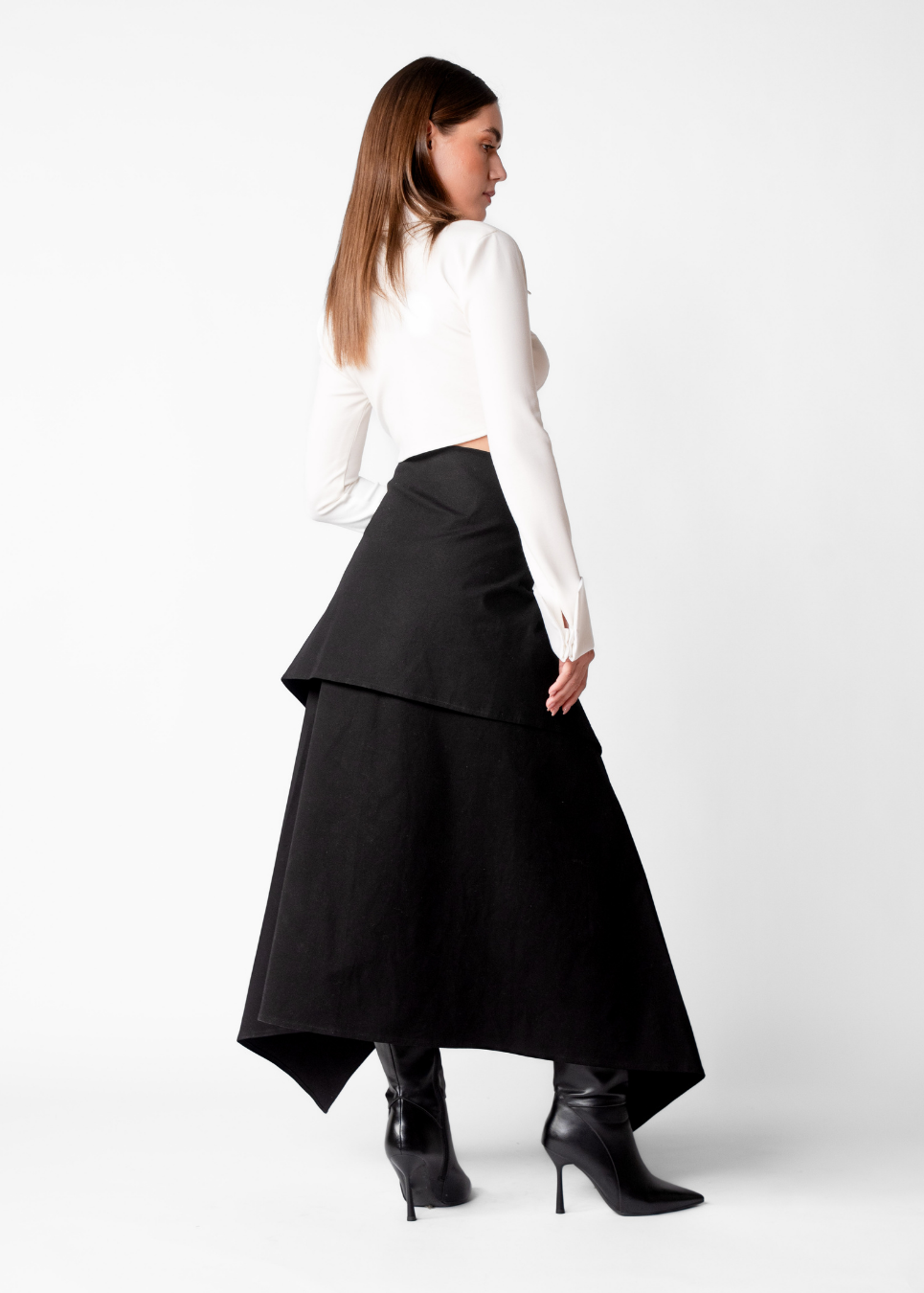 A woman shown from the back wearing a white long-sleeve crop top, a black high-low tiered skirt, and black pointed-toe heeled boots, photographed against a clean white studio background.