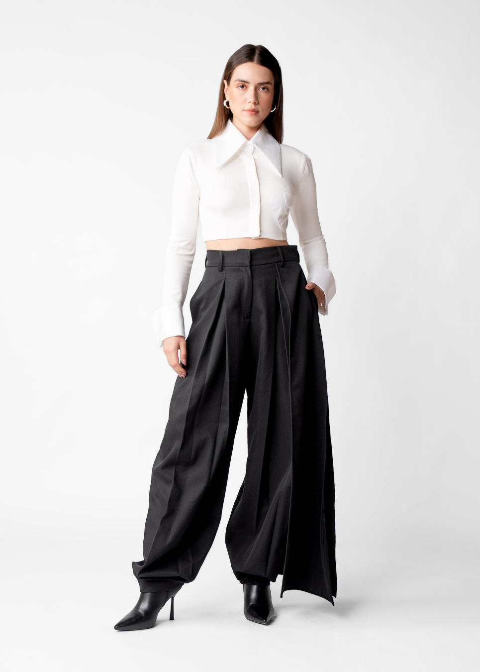 A woman modeling a white long-sleeve cropped button-up shirt paired with high-waisted black wide-leg trousers and black pointed-toe ankle boots, photographed against a seamless light gray studio background.
