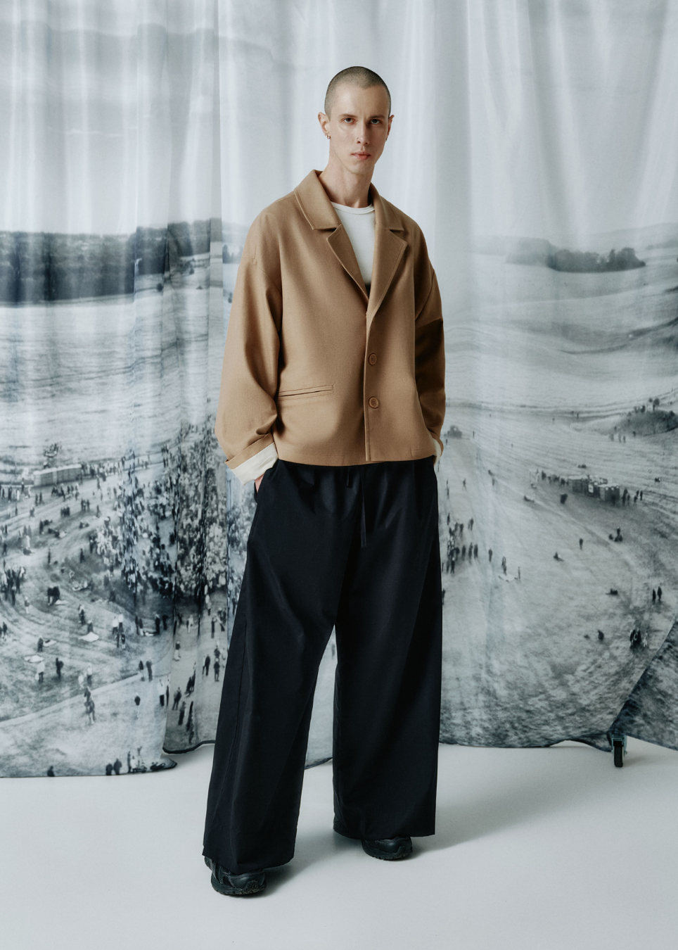 A model stands in a studio wearing a camel-colored oversized blazer over a white shirt and black wide-leg trousers, posed in front of a large black-and-white landscape photograph backdrop.