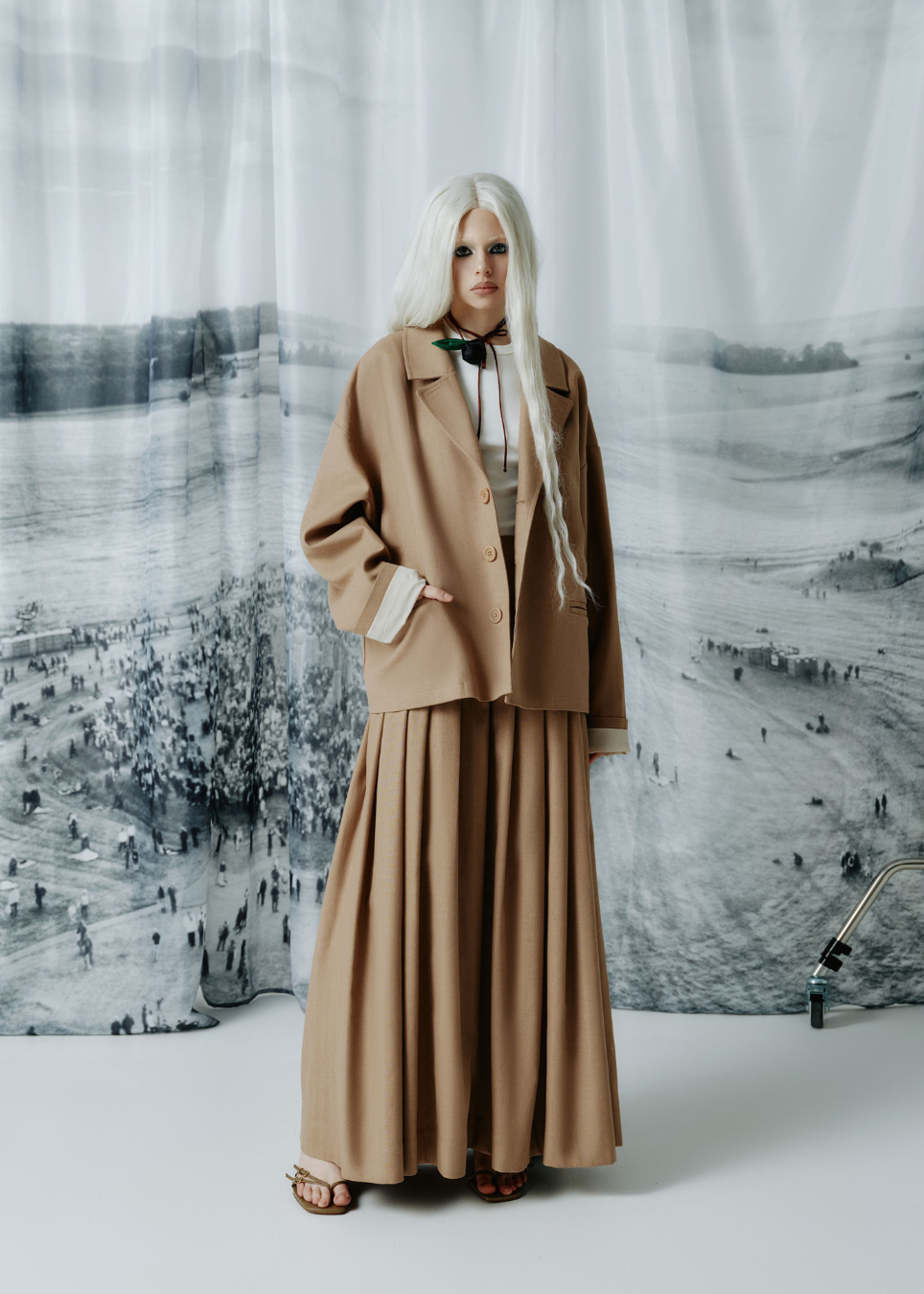 A model with long platinum blonde hair stands in front of a large black and white scenic backdrop, wearing a matching beige two-piece suit consisting of a cropped blazer and a long pleated skirt.