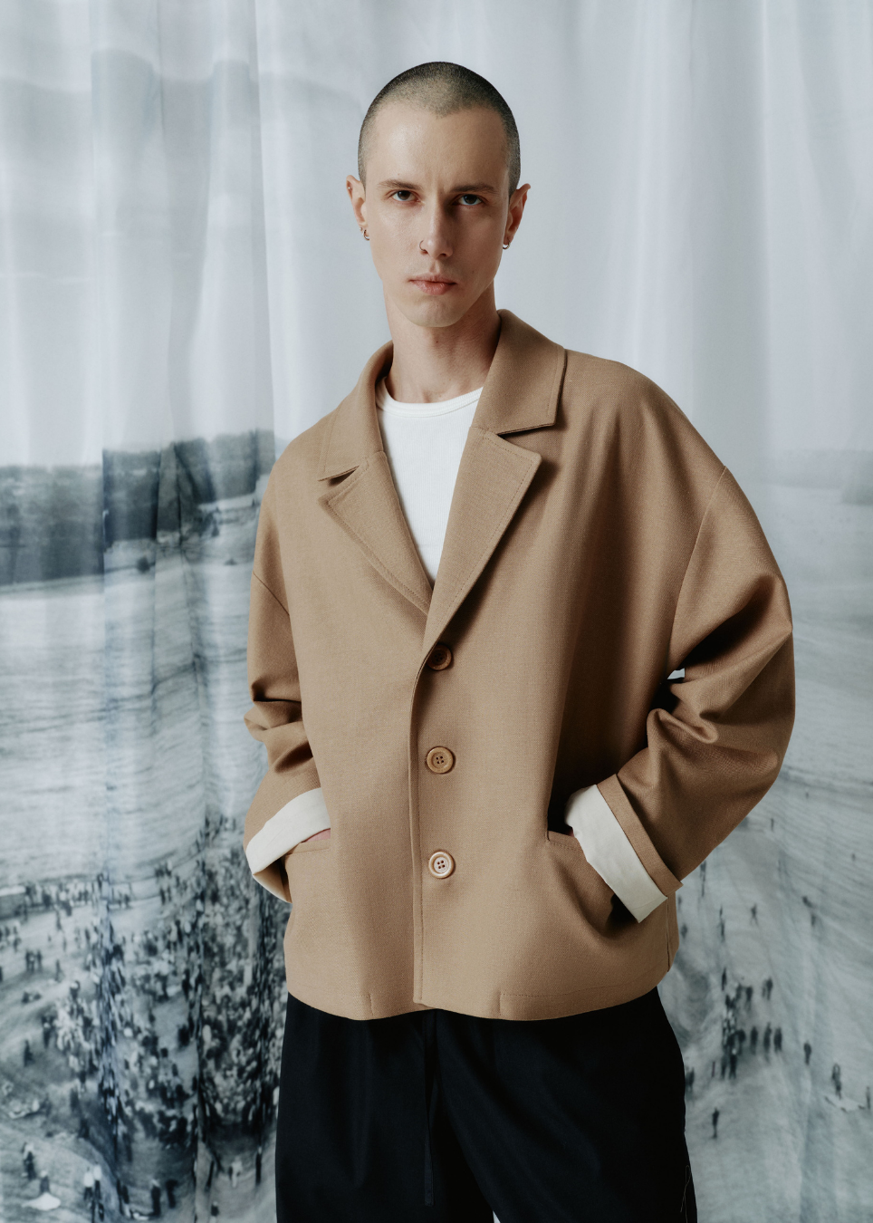 A model with a shaved head wearing a camel-colored oversized blazer with contrasting white cuffs, layered over a white t-shirt and dark trousers, standing in front of a soft-focus gray and white background.