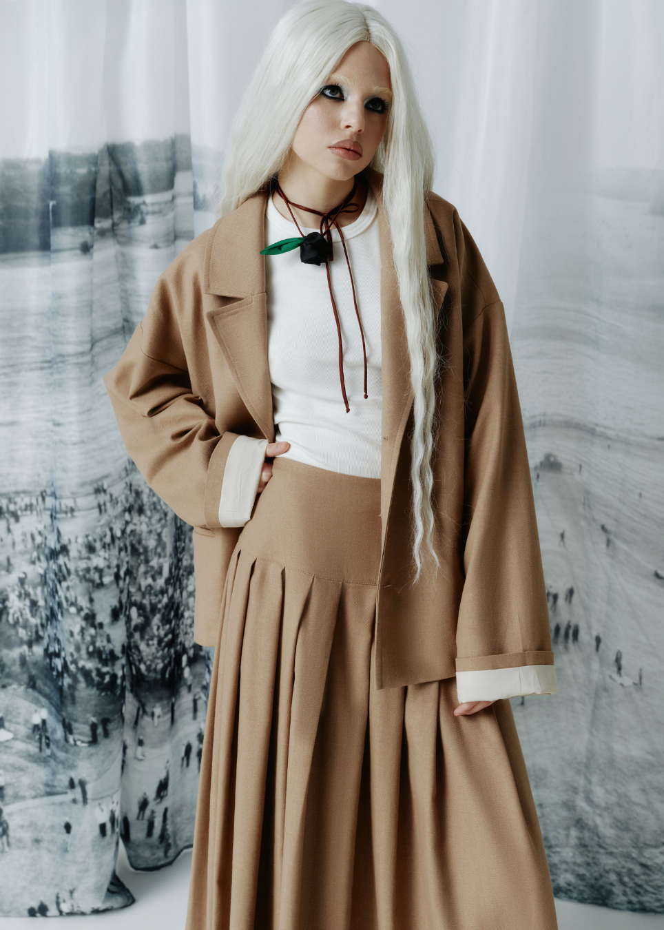 A model with long platinum blonde hair wearing a camel brown coat over a white top and matching pleated skirt, styled with a brown cord necklace, posed in front of a gray and white scenic backdrop.