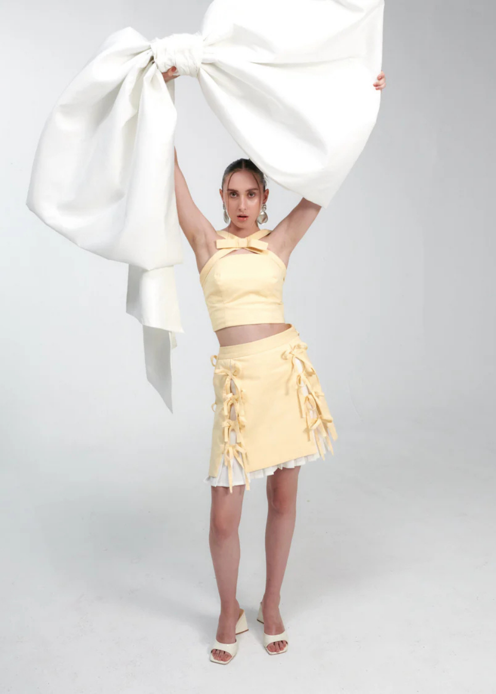 A model stands against a light gray studio background wearing a pale yellow halter crop top and matching ruffled mini skirt, while holding a large white fabric bow overhead.