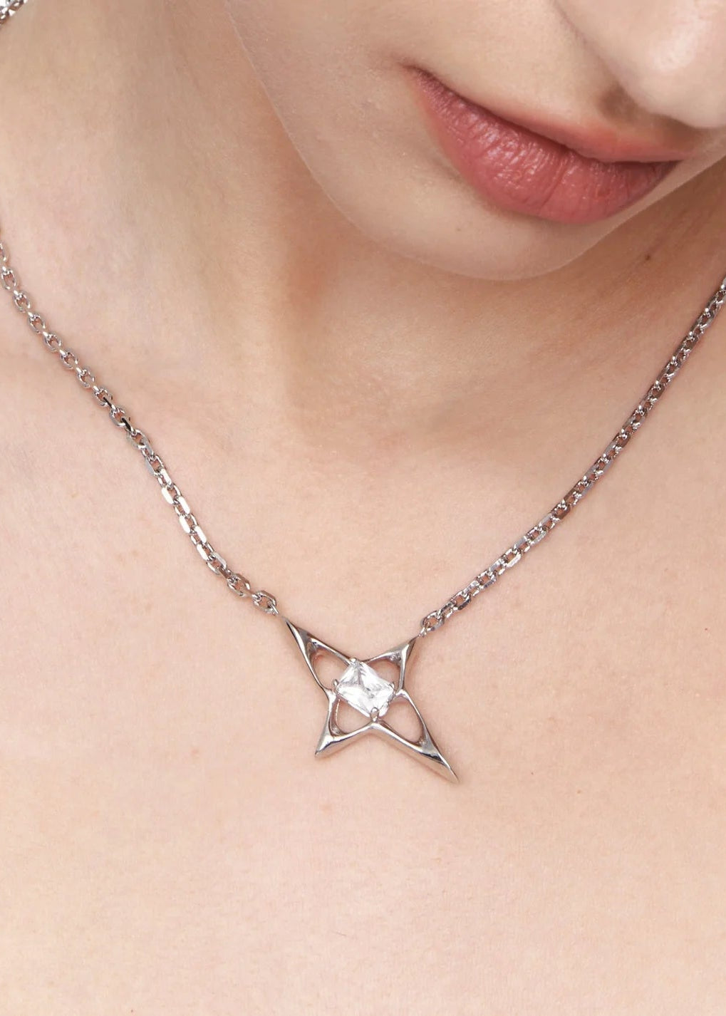 A close-up of a model's neck and chest wearing a silver chain necklace with a geometric star-like pendant featuring a clear central gemstone.